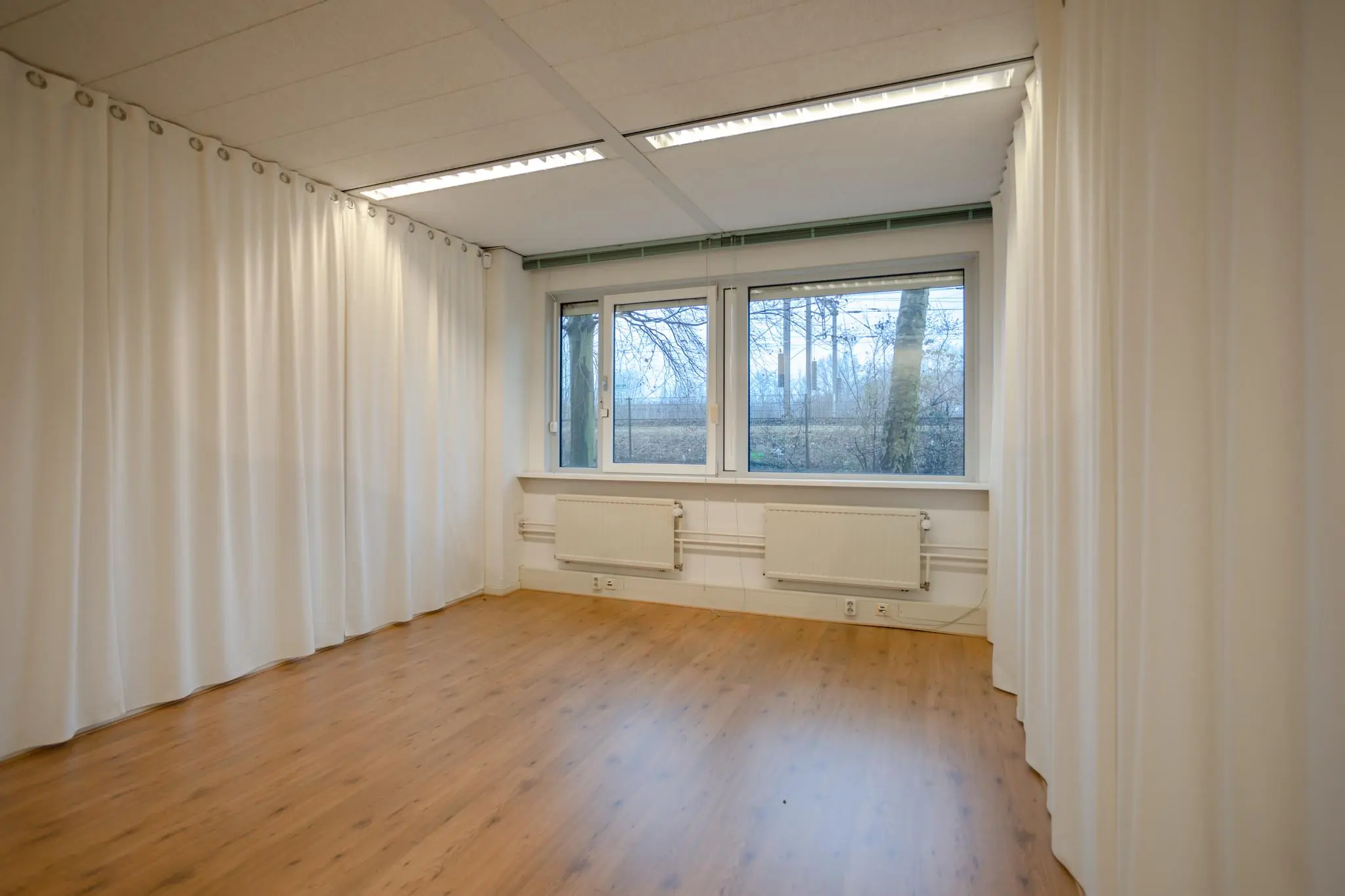 Lichte, lege kamer met houten vloer, witte gordijnen en grote ramen aan de Christiaan Krammlaan.